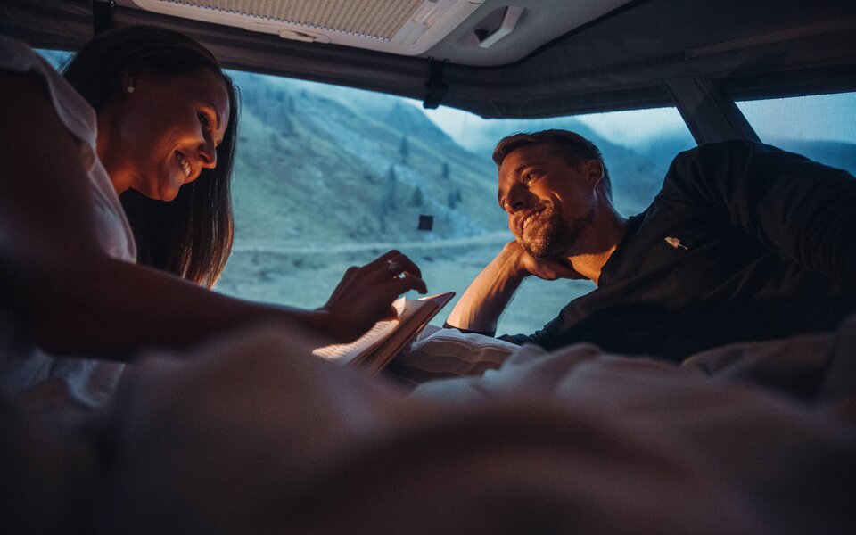Un couple est allongé dans le lit d’un camping-car Carado et contemple le paysage montagneux au crépuscule