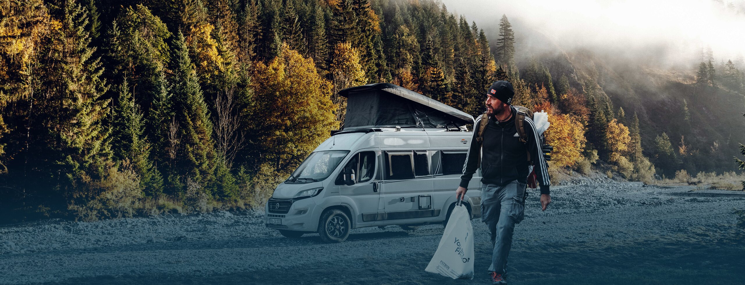 Person standing in front of a Carado camper van with pop-up roof on a gravel parking area at the edge of the forest