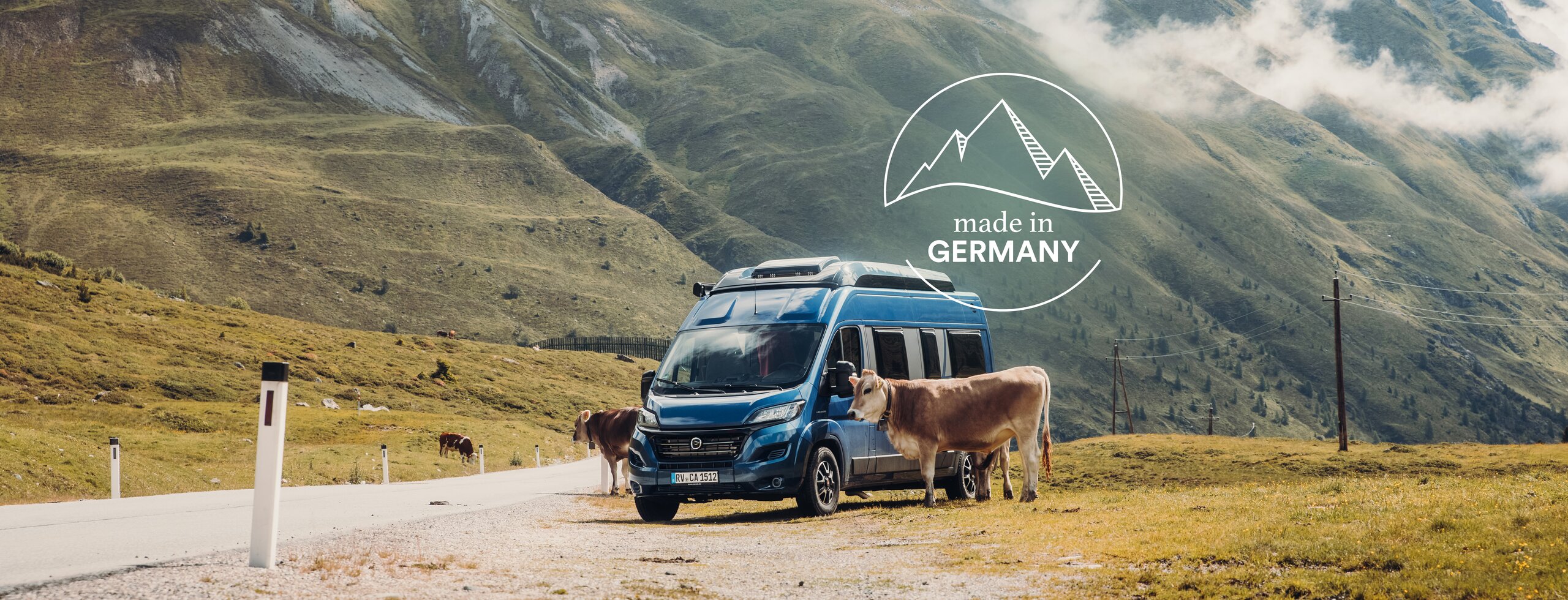 Blue Carado camper van on a mountain pass beside cows, with green Alpine scenery in the background