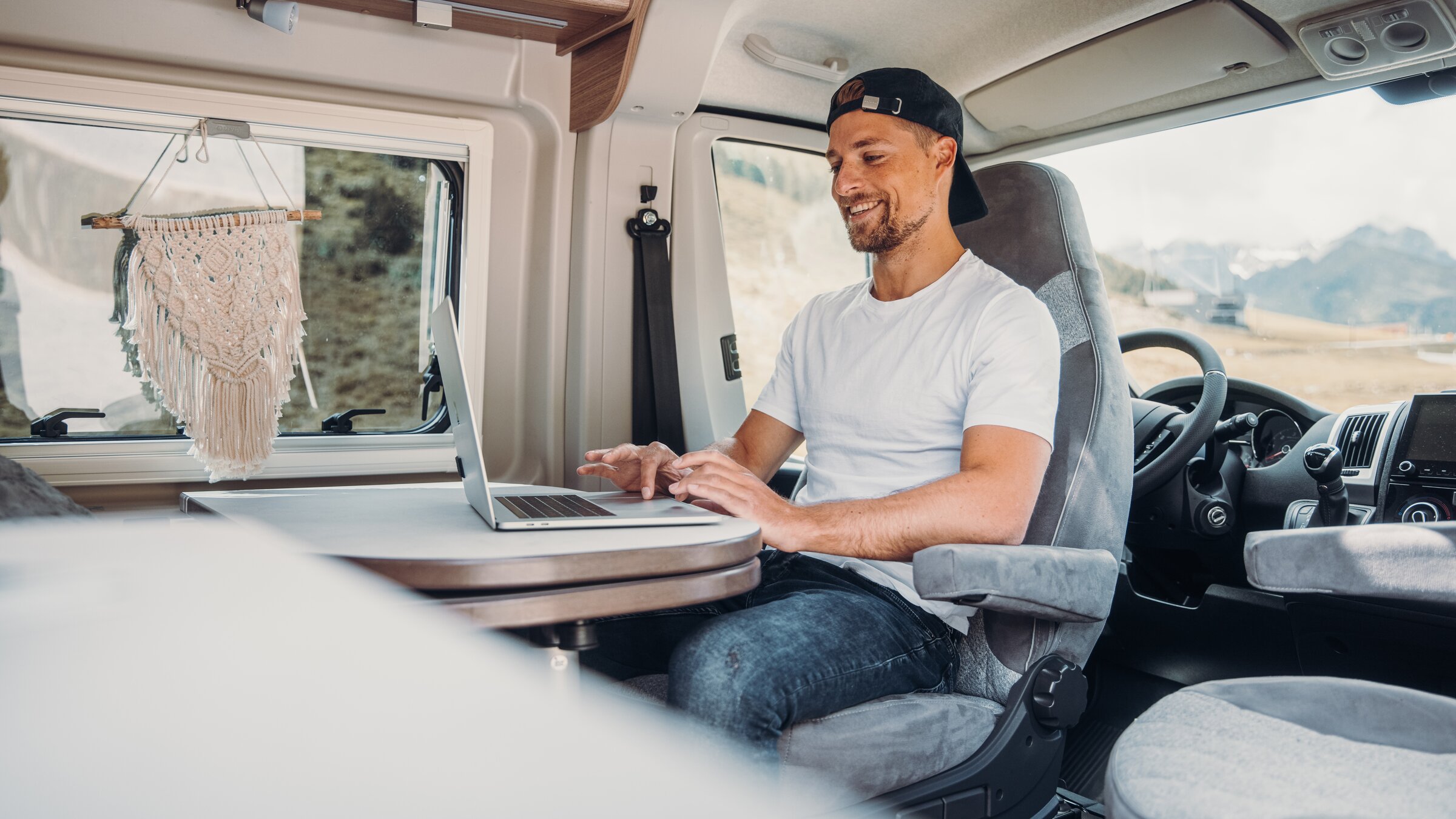 Man working on a laptop at the dinette in the Carado motorhome, with the cab and interior windows visible