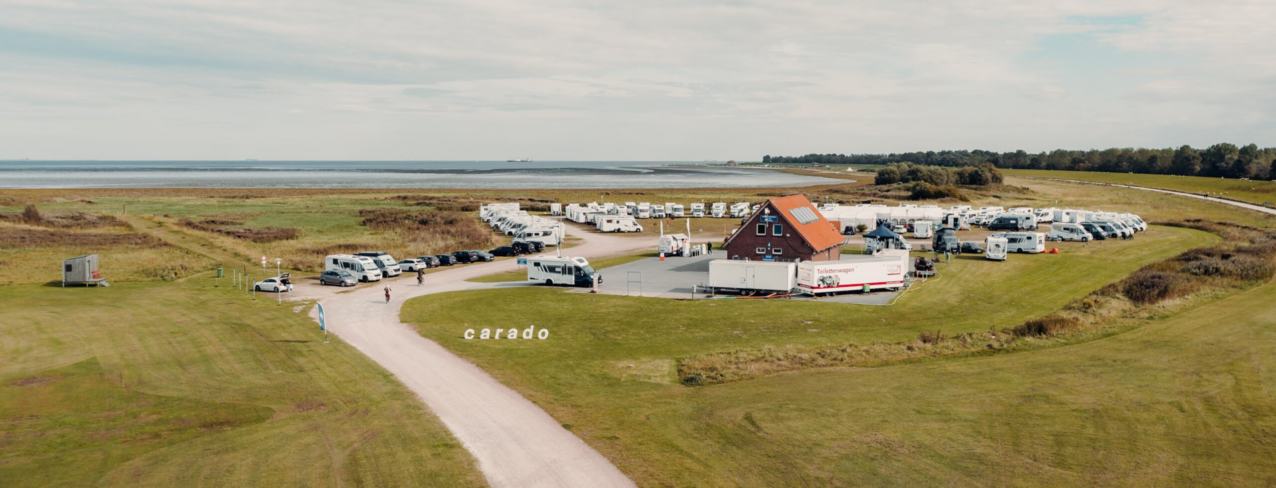 Aerial view of a Carado motorhome meet-up at a coastal campsite with many vehicles and a building by the water