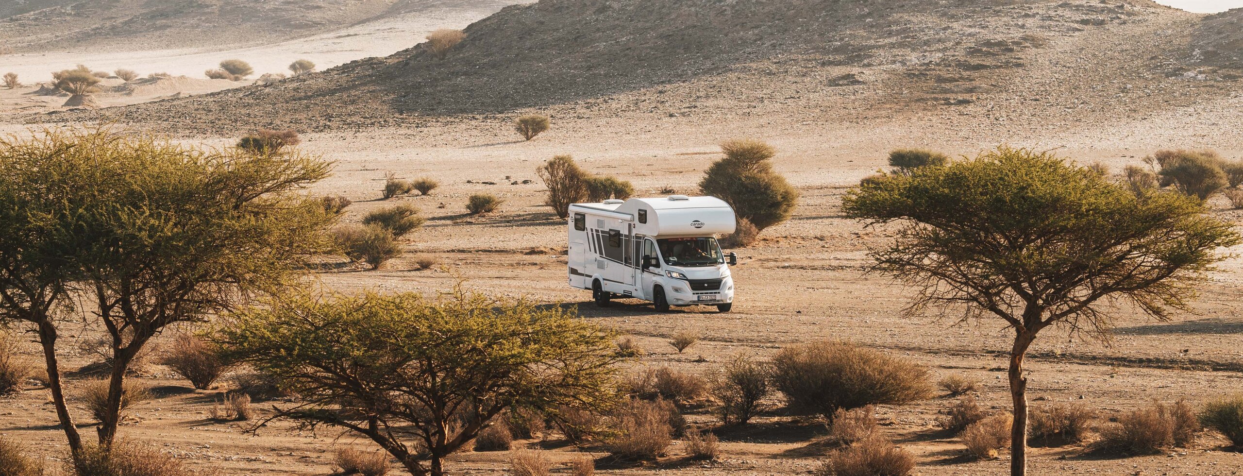 Carado camper in een kaal woestijnlandschap met bomen en rotsachtige heuvels