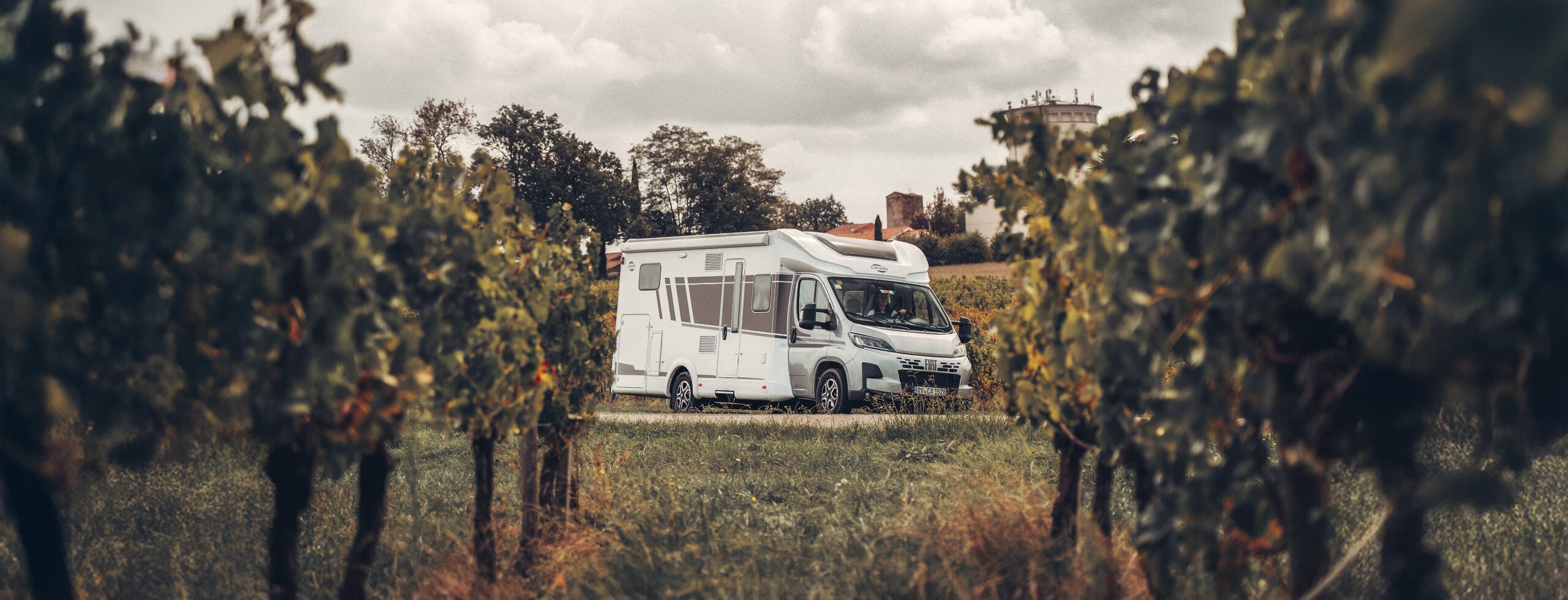 Camping-car Carado au milieu des vignes sur un chemin de terre, vue de face et de côté sous un ciel nuageux