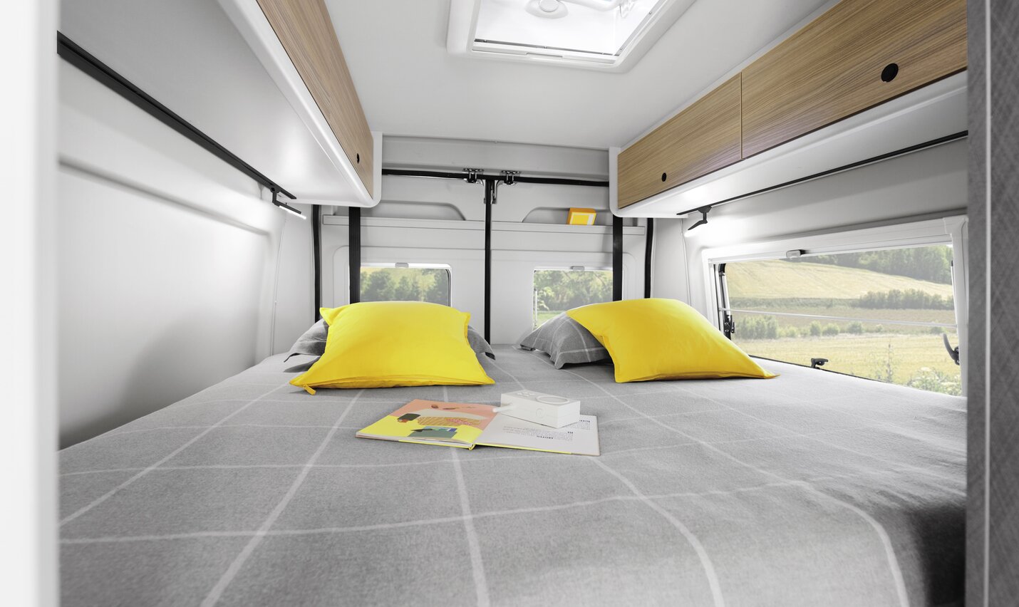 Sleeping area of a Campervan Fiat Plus with double bed, gray bedding, striped pillow, and view through window of green landscape.