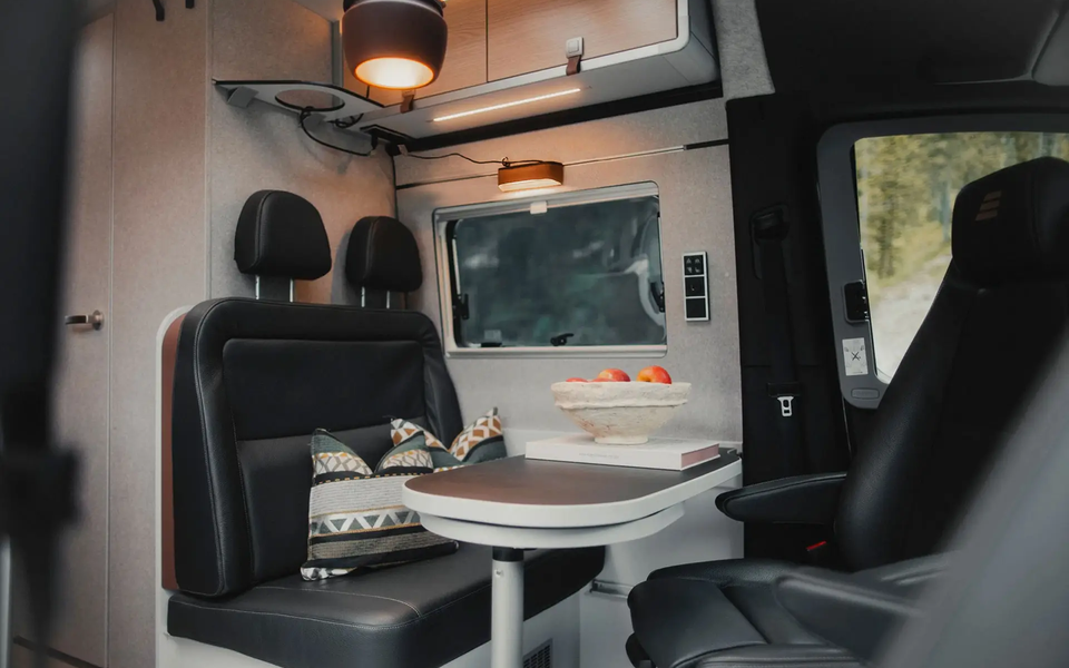Living area in a modern camper van, set table with seating area with dark leather, the two driver's seats are turned towards the table, above the seating area are roof storage cupboards and a brown pendant lamp
