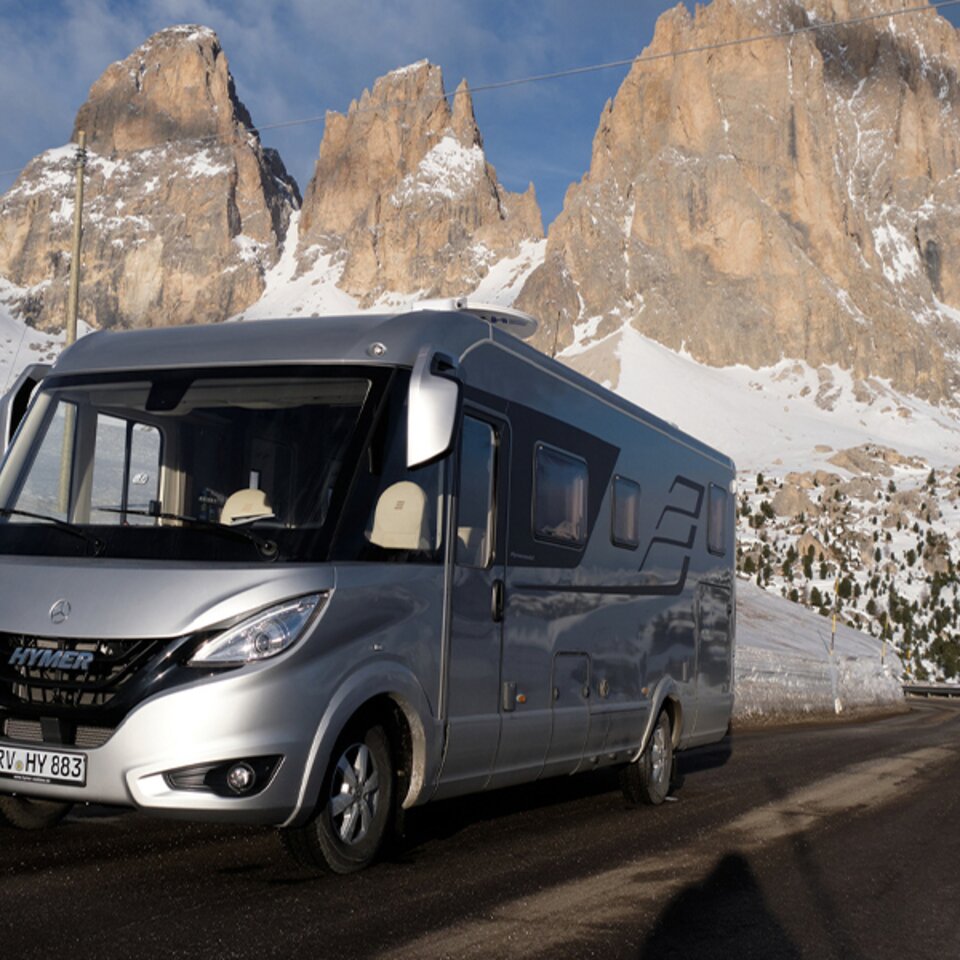 Un HYMER B-Klasse MasterLine I argenté est stationné par temps ensoleillé sur une route asphaltée, avec des montagnes enneigées en arrière-plan.