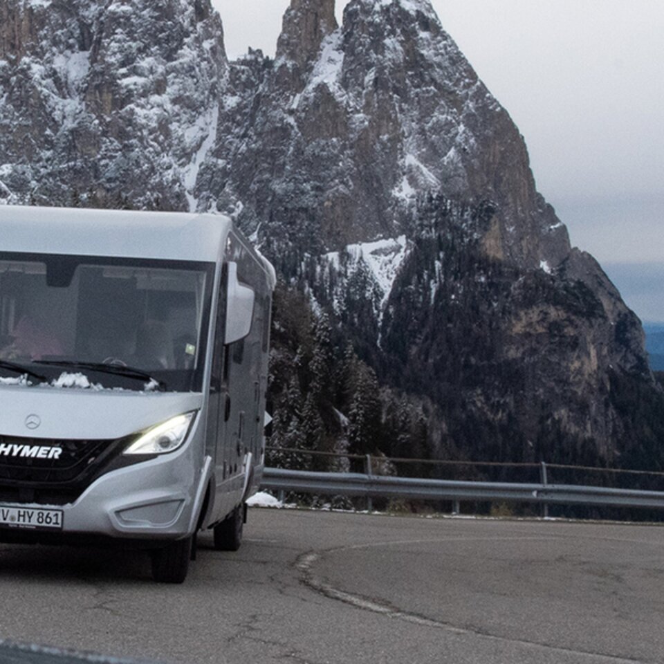 Un HYMER B-Klasse ModernComfort I gris est garé avec des phares allumés dans un virage d'une route de montagne, avec de la neige sur les montagnes derrière.