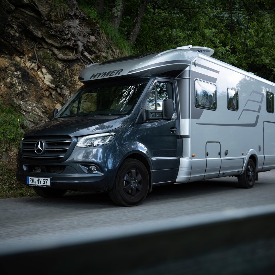 Le HYMER B-Klasse MasterLine T avec cabine sombre et carrosserie grise est stationné sur une route avec un talus rocheux et des arbres verts sur le bord.