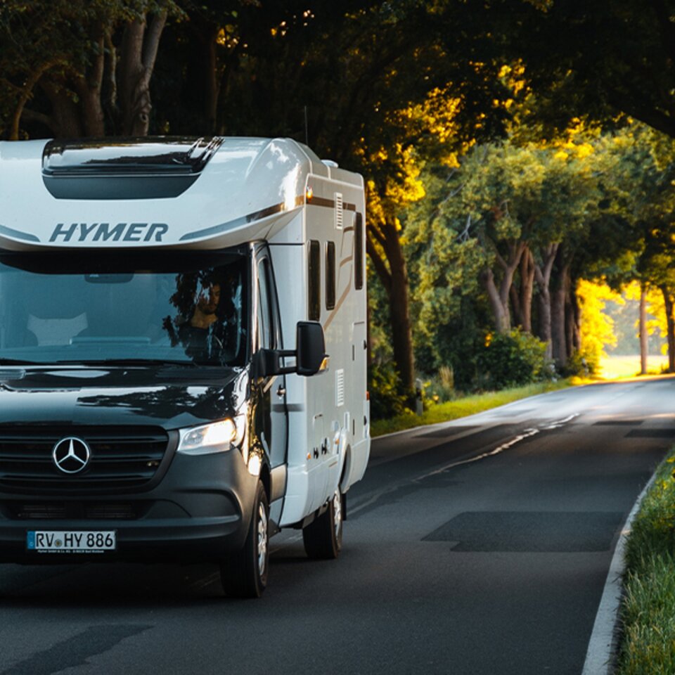 Un HYMER B-Klasse ModernComfort T noir et blanc roule par temps ensoleillé avec des phares allumés à travers une allée bordée d'arbres verdoyants.