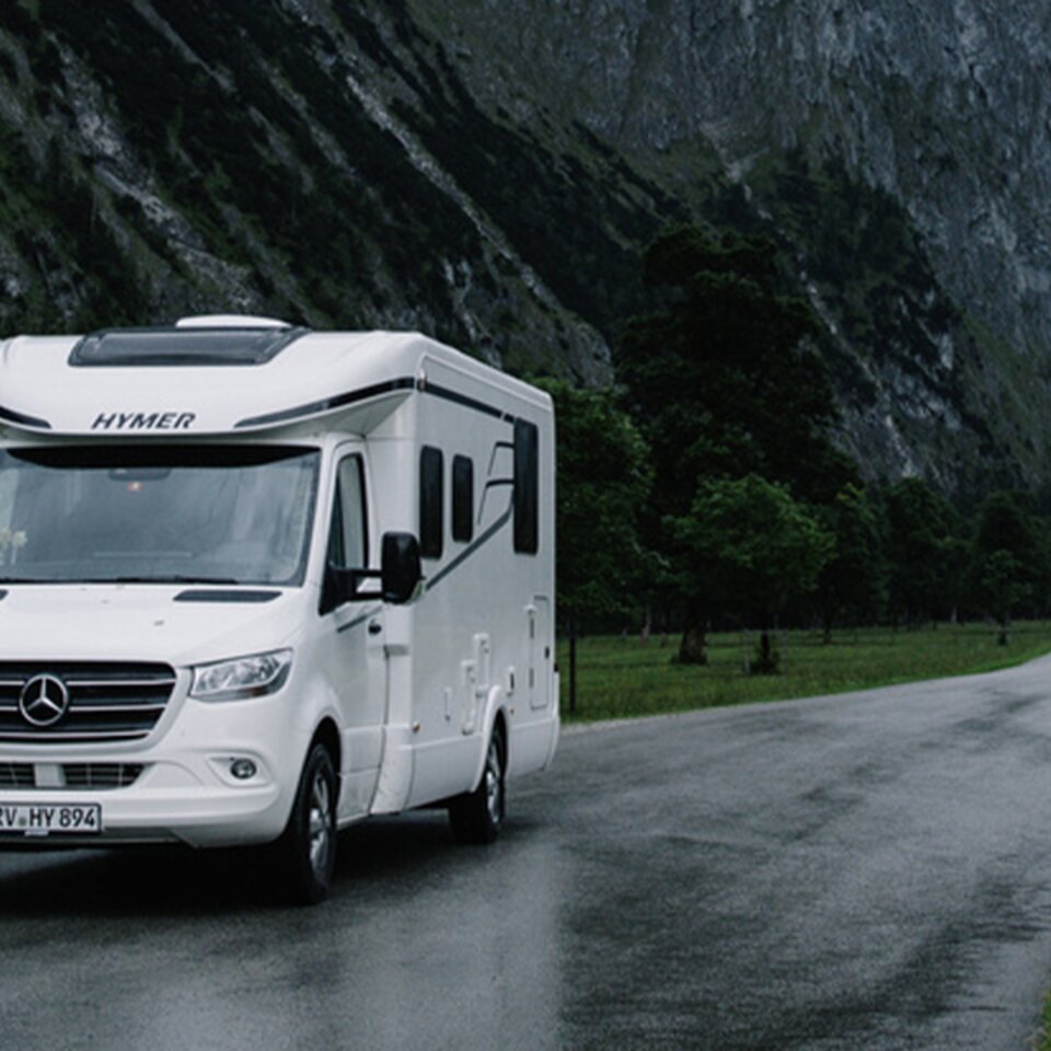 Un HYMER Tramp S blanc est stationné sur une route étroite et humide dans un paysage montagneux, avec des montagnes, des arbres et des prairies en arrière-plan.
