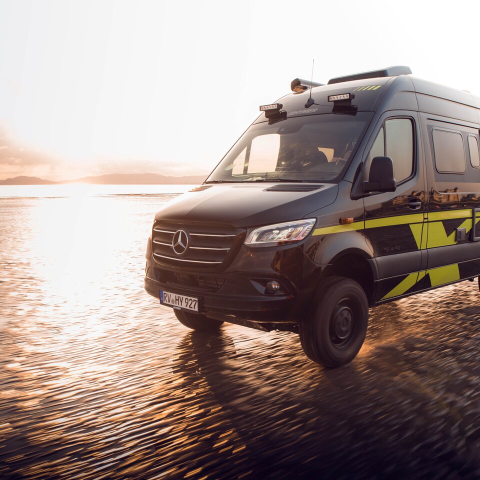 Das Mercedes Allrad-Wohnmobil HYMER Grand Canyon S fährt schnell auf einem sonnigen, nassen Strand. Die Umgebung ist aufgrund der Dynamik unscharf.