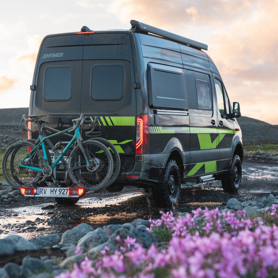 Das Sprinter-4x4-Wohnmobil HYMER Grand Canyon S CrossOver steht auf einem unbefestigten Weg mit Blumen und Gras, auf dem Fahrradträger stehen Räder.