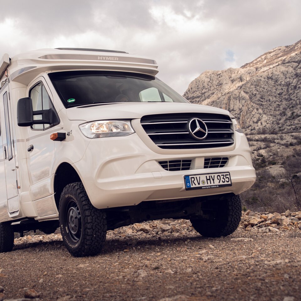 Das weiße Mercedes Allrad-Wohnmobil HYMER ML-T steht auf einer ansteigenden Steinstraße mit felsigen Hügeln und leicht wolkigem Himmel im Hintergrund.
