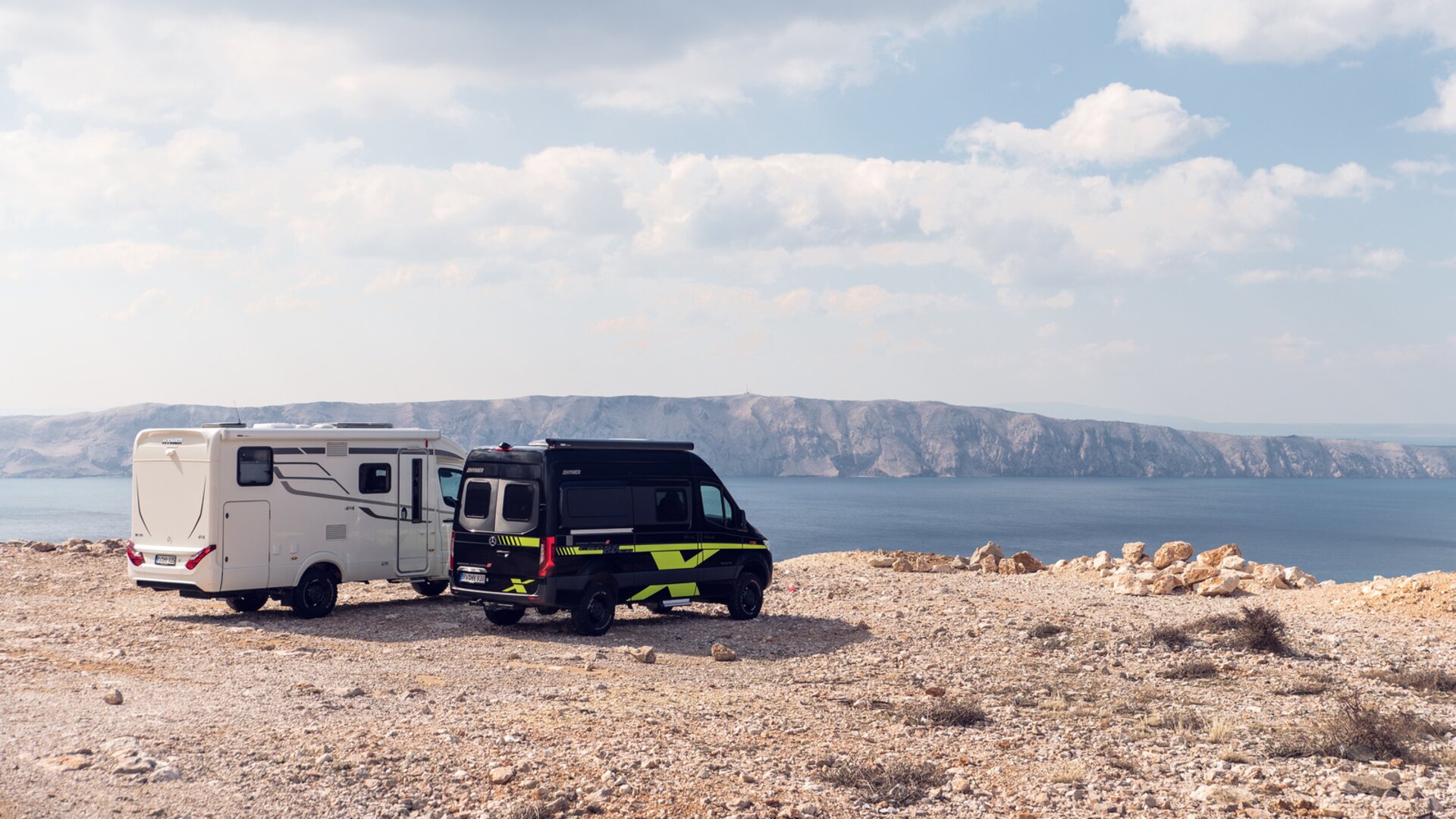 Zwei Offroad-Wohnmobile in Weiß sowie Schwarz-Grün stehen auf einer sonnenbeschienenen Ebene über dem Meer, im Hintergrund sind Klippen zu sehen.