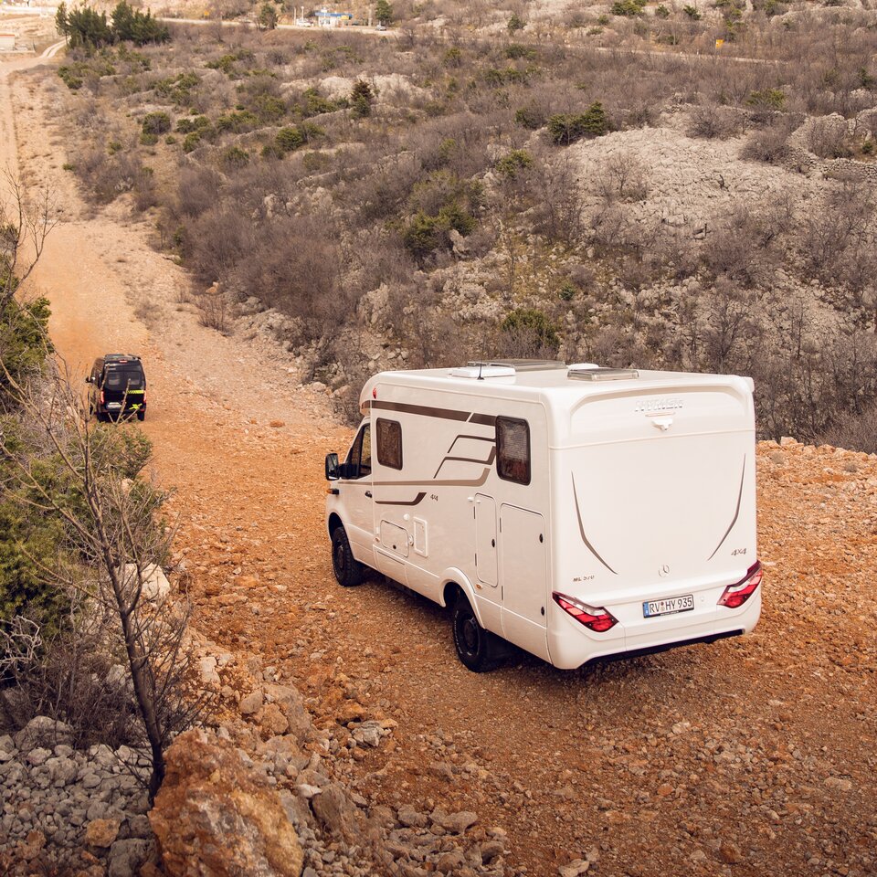 Zwei 4x4 Offroad-Wohnmobile von HYMER mit Mercedes-Triebkopf fahren auf einer hügeligen Straße durch unbefestigtes, steiniges Gelände mit Sträuchern. 