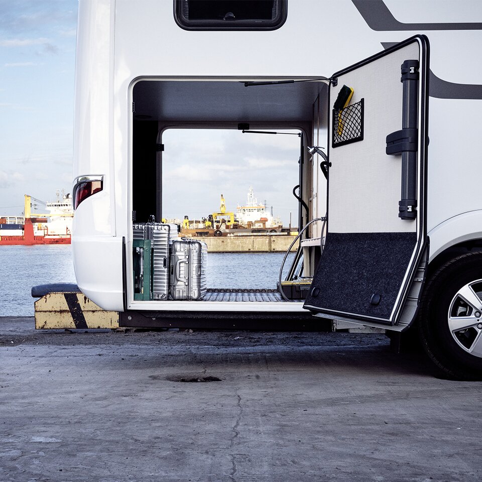 Dans le garage ouvert du HYMER B-Klasse Master Line I, un camping-car avec tête motrice Mercedes, il y a des valises. En arrière-plan, on voit un port.