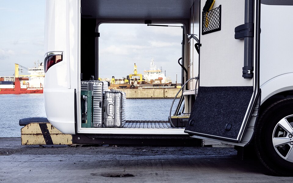 There are suitcases in the open garage of the HYMER B-Class Master Line I, a motorhome with a Mercedes power unit. A harbour can be seen in the background.