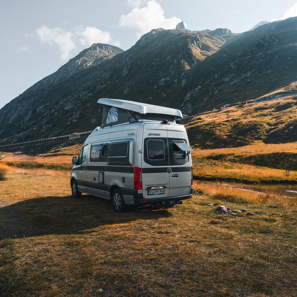 Le HYMER Grand Canyon S est vu légèrement de derrière, à un angle. Il est légèrement tourné devant un panorama de montagne sur un pré.