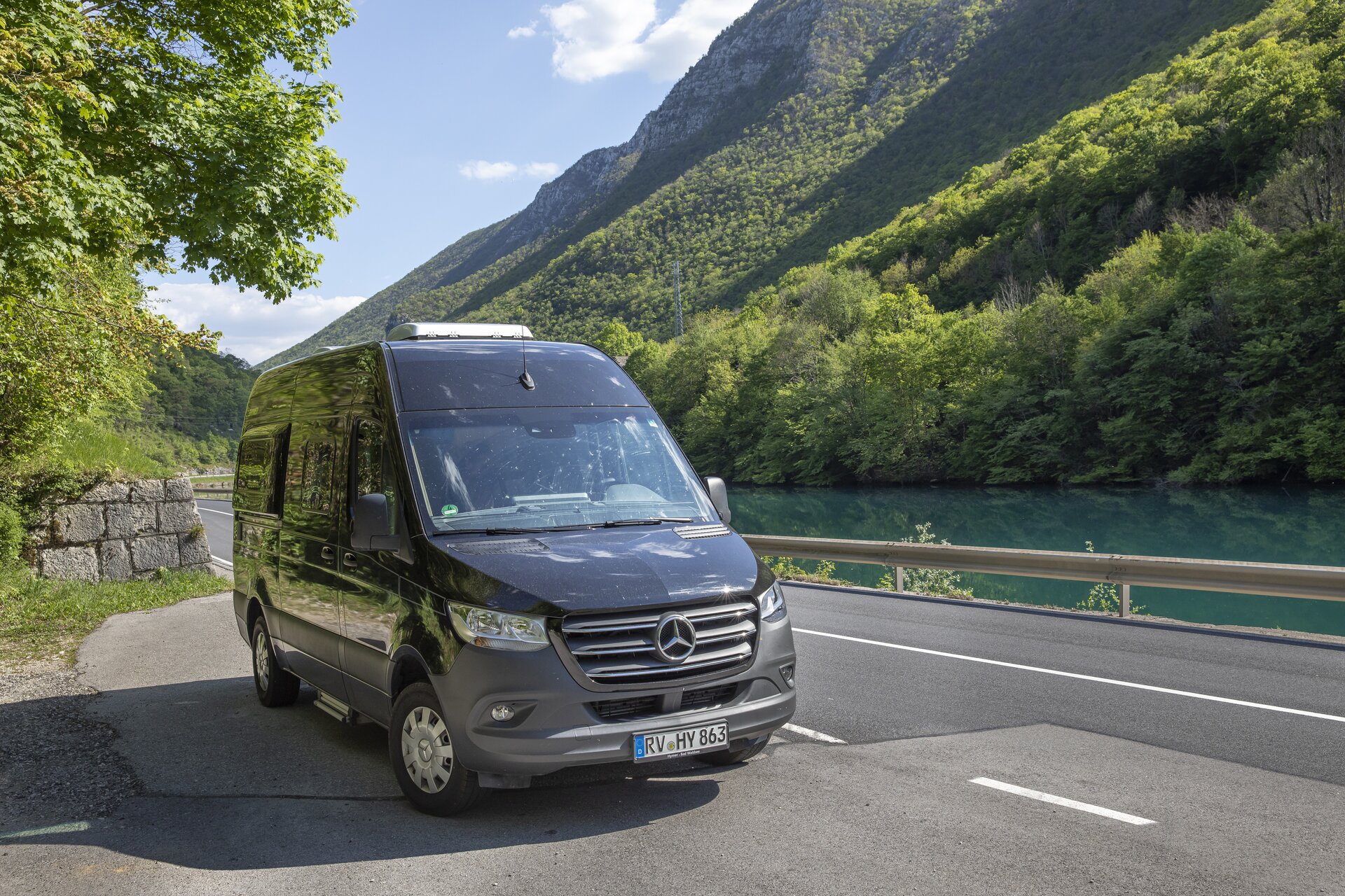 The black HYMER Grand Canyon S Mercedes camper van is parked on a road next to a river in a mountainous landscape, with trees growing on the mountains.