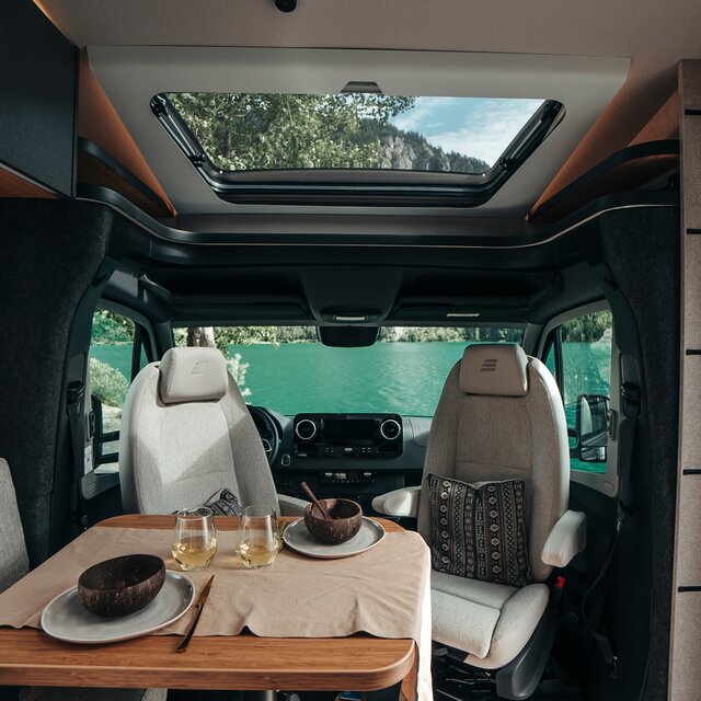La table du HYMER ML-T dans le monde du style Native Bamboo est dressée avec de la vaisselle et du vin blanc. Derrière se trouve la cabine de conduite avec vue sur la nature.
