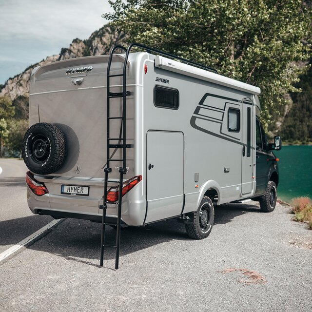 Le HYMER ML-T est vu de biais arrière. Il est légèrement tourné vers un lac de montagne et l'échelle de toit est déployée.