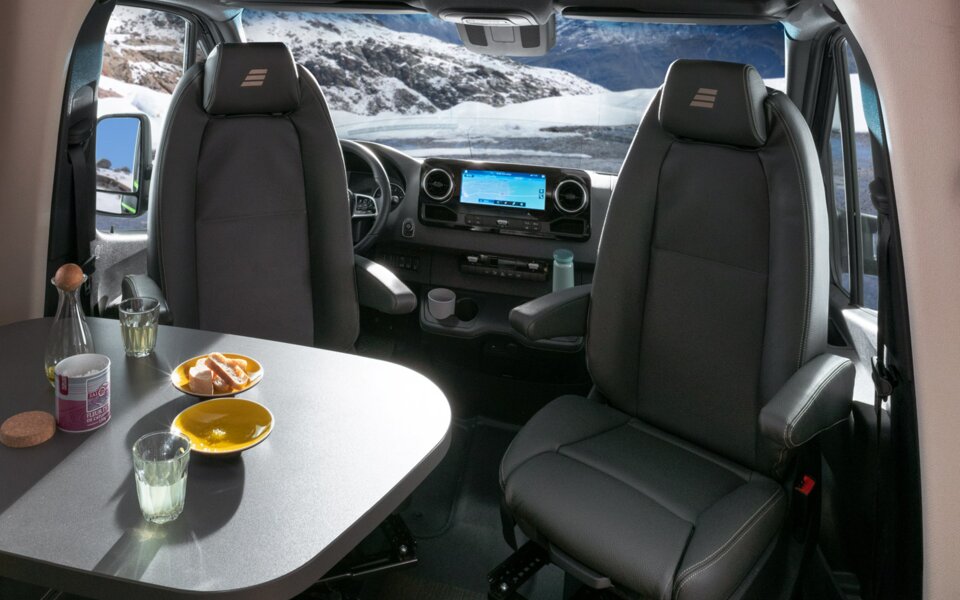 Living space in the HYMER ML-T CrossOver: dark leather seating area, laid table, panoramic roof vent, driver's seats and driver's cab