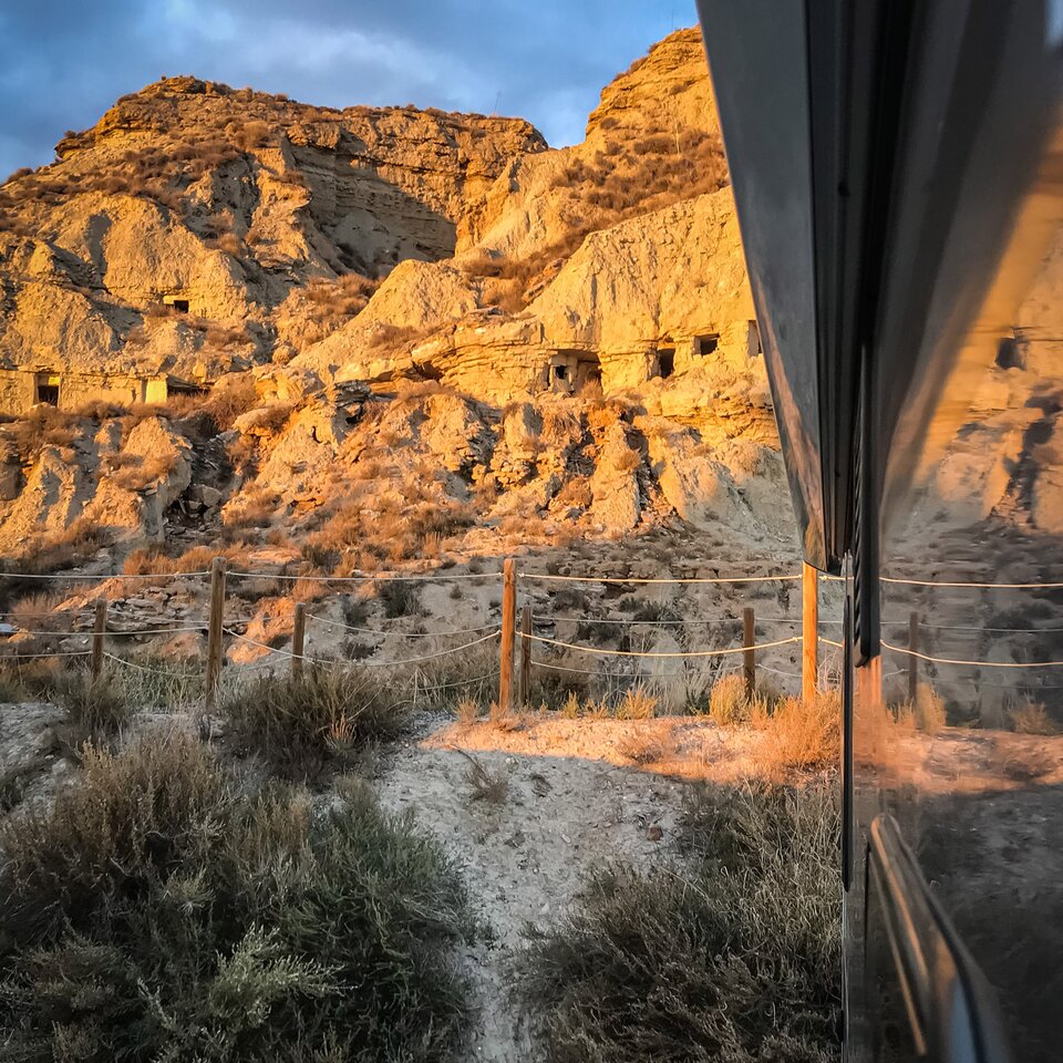 View from the motorhome into the Pyrenees landscape, the wall of the motorhome can be seen to the side, bushes and stony, sunlit hills behind.