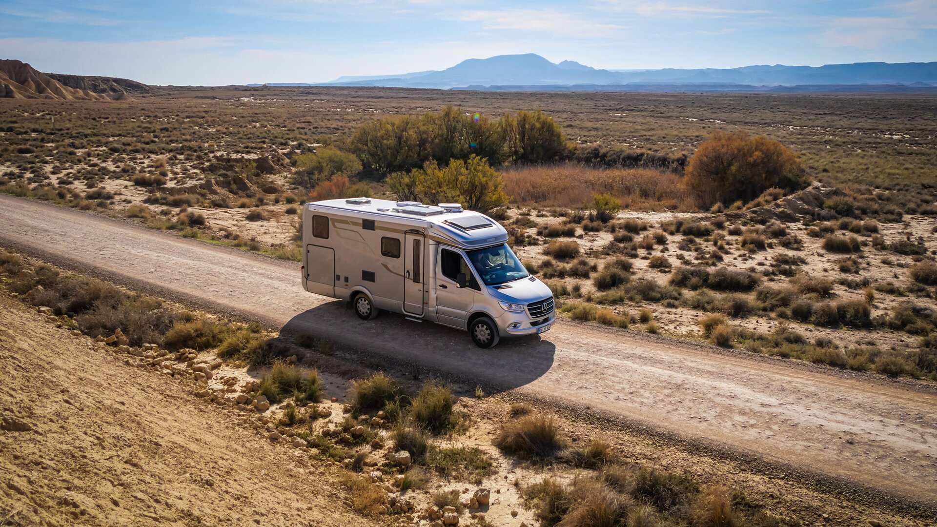 Picture of a journey through the Pyrenees in a motorhome. The silver vehicle is parked on a road in barren surroundings with grass, sand and mountains.